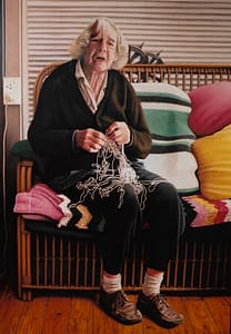 An oil painting by Josonia Palaitis depicting her elderly mother sitting on a couch with coloured knitted blankets behind her holding a messy ball of string and looking anguished