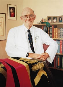 Portrait painting by Josonia Palaitis depicting Dr David Tiller sitting in front of a bookshelf with his hands together looking relaxed and wearing his white physician coat