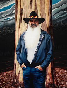 An oil painting by Josonia Palaitis depicting Patrick Dodson standing in front of a gum tree trunk wesaring jeans with his hands in his pockets and an open blue jacket that is filled by his huge white beard and he's wearing a black hat with black, red and yellow stripes in recognition of his Aboriginal ancestry