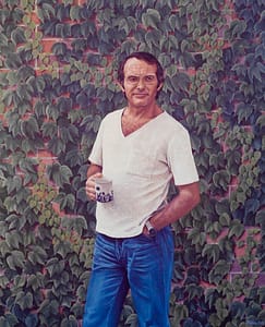 Oil painting by Josonia Palaitis depicting musician Adrian Dangerfield standing in front of a brick wall covered with vine leaves holding a mug of tea with the other hand in his pocket wearing jeans and a white t-shirt