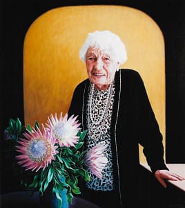 Oil painting by Josonia Palaitis depicting her aunty at the age of 100 standing in front of a yellow background wearing a black cardigan and bead necklaces with pink Australian wildflowers in the foreground
