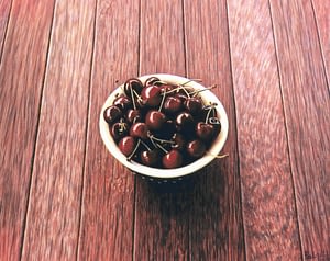 Photorealistic painting by Josonia Palaitis depicting a full bowl of cherries placed on a wooden floor