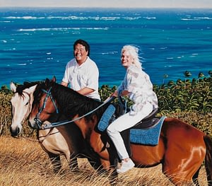 Oil painting by Josonia Palaitis featuring a man and a woman both on horseback in front of the ocean on a windy yet sunny day, both smiling and looking at the viewer
